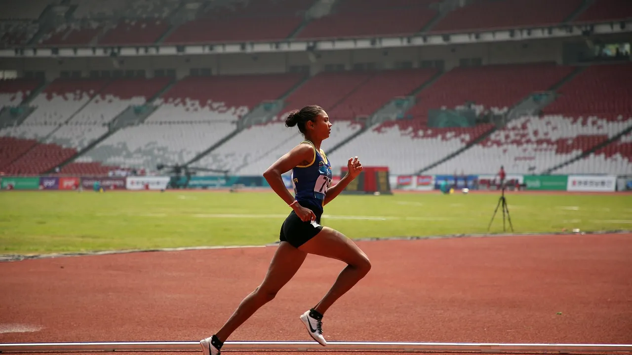 Una corredora con equipo de competición corre de perfil por una pista de atletismo roja, frente a un estadio vacío, a plena luz del día.